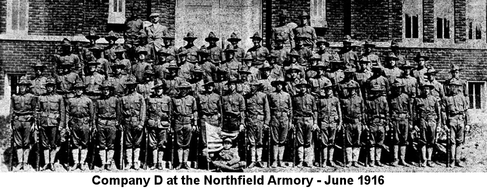 Black and white newspaper photo of dozens of men in World War I army uniforms and campaign hats standing in rows before a large brick armory building.